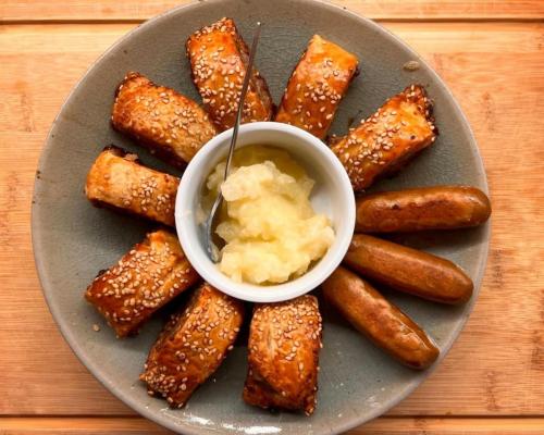 Gluten-free sausage rolls and vegan sausages around a small pot of apple sauce.